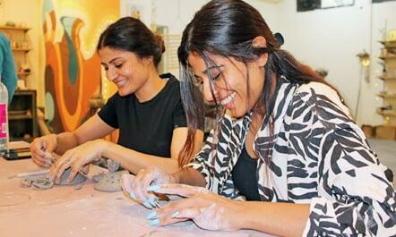 Two girl participants enjoying handbuilding at Sona Pottery. Both are similing and immersed in shaping the clay